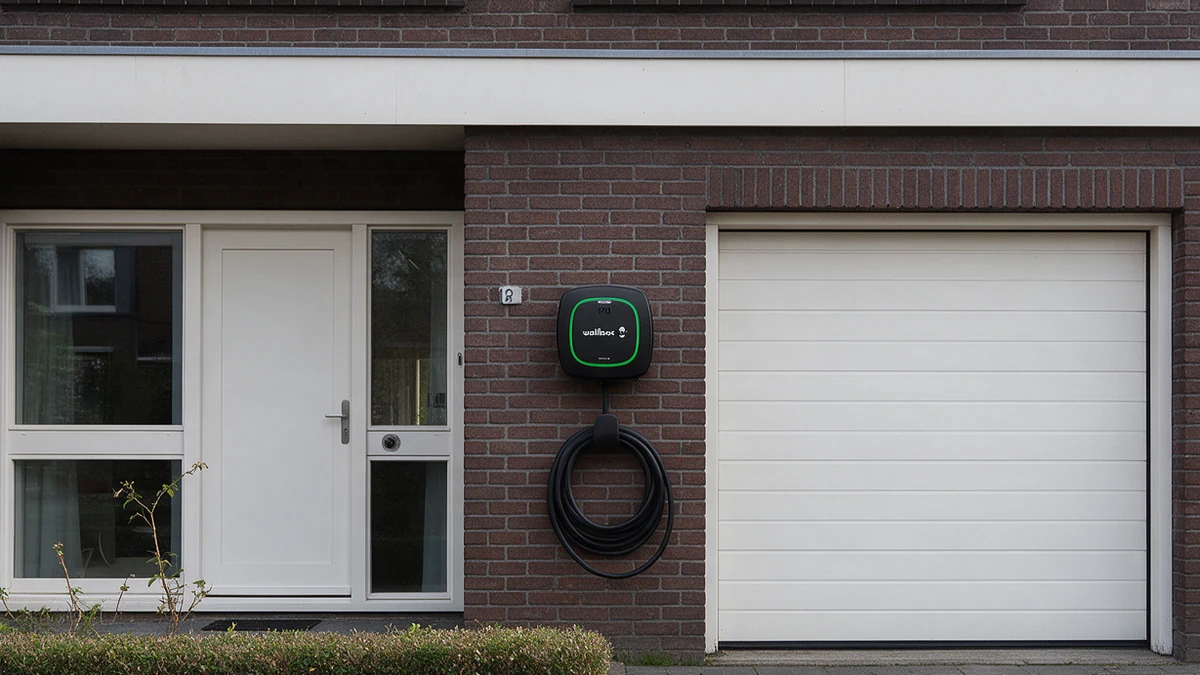 Zwarte Wallbox Pulsar Plus laadpaal met groene lichtring en opgerolde kabel naast de witte garagedeur van een Nederlandse rijtjeswoning van donkerrode baksteen, met een lage haag.