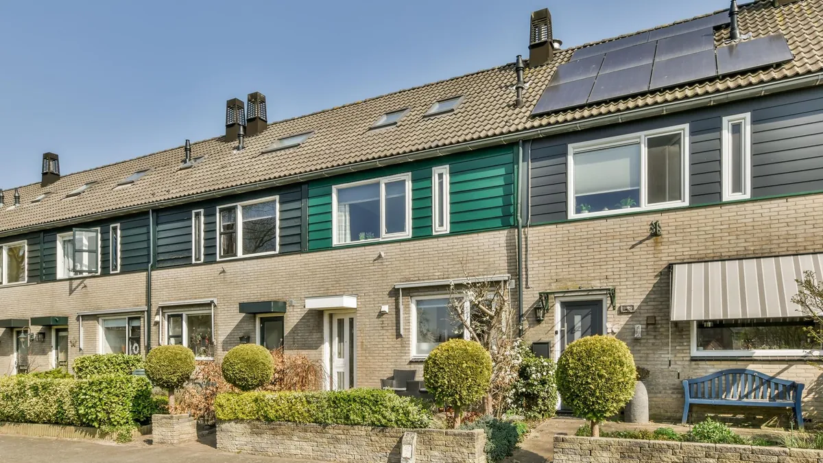 Rij Nederlandse rijtjeshuizen van gele baksteen met donkere en groene gevelbeplanking, zonnepanelen op het dak en voortuinen met bolvormige struiken.
