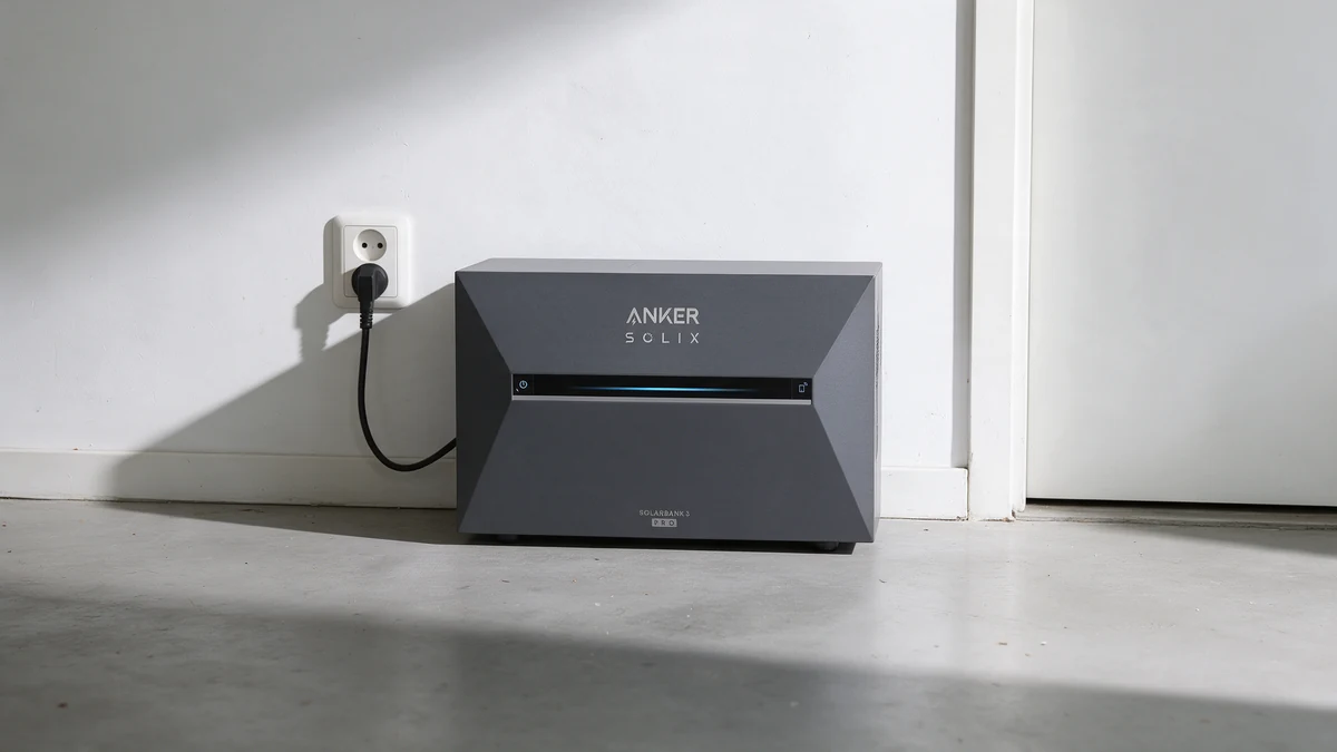 Grijze Anker SOLIX Solarbank 3 thuisbatterij op de betonvloer tegen de witte muur van een berging in een Nederlandse rijtjeswoning, met de stekker in een wandcontactdoos en een witte deur ernaast.
